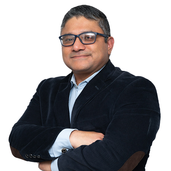 Un hombre con gafas y pelo corto y oscuro, de pie, con los brazos cruzados, vestido con americana oscura y camisa azul claro, sobre un fondo blanco liso, encarna la profesionalidad en fisioterapia y ortopedia deportiva.