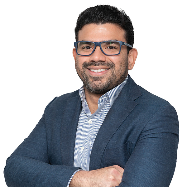 Un hombre moreno con gafas y sonrisa confiada, vestido con americana azul y camisa a rayas, de pie con brazos cruzados sobre un fondo blanco, transmitiendo profesionalismo en fisioterapia y ortopedia deportiva.