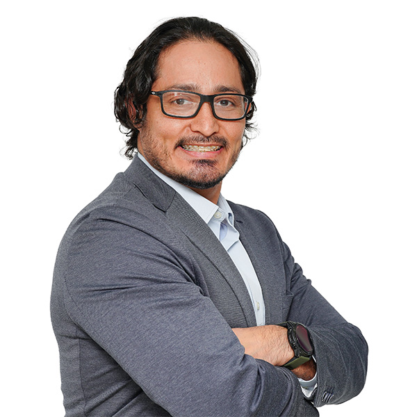 Hombre con pelo oscuro y gafas, vestido con americana gris y camisa clara, sonriendo con los brazos cruzados, de pie sobre un fondo blanco liso-especializado en ortopedia deportiva y prevención de lesiones.