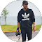 Persona en exteriores, con sudadera y gorra negras, sostiene una cámara cerca de una carretera con agua y hierba de fondo, capturando momentos relacionados con fisioterapia o lesiones, en un entorno natural y tranquilo.