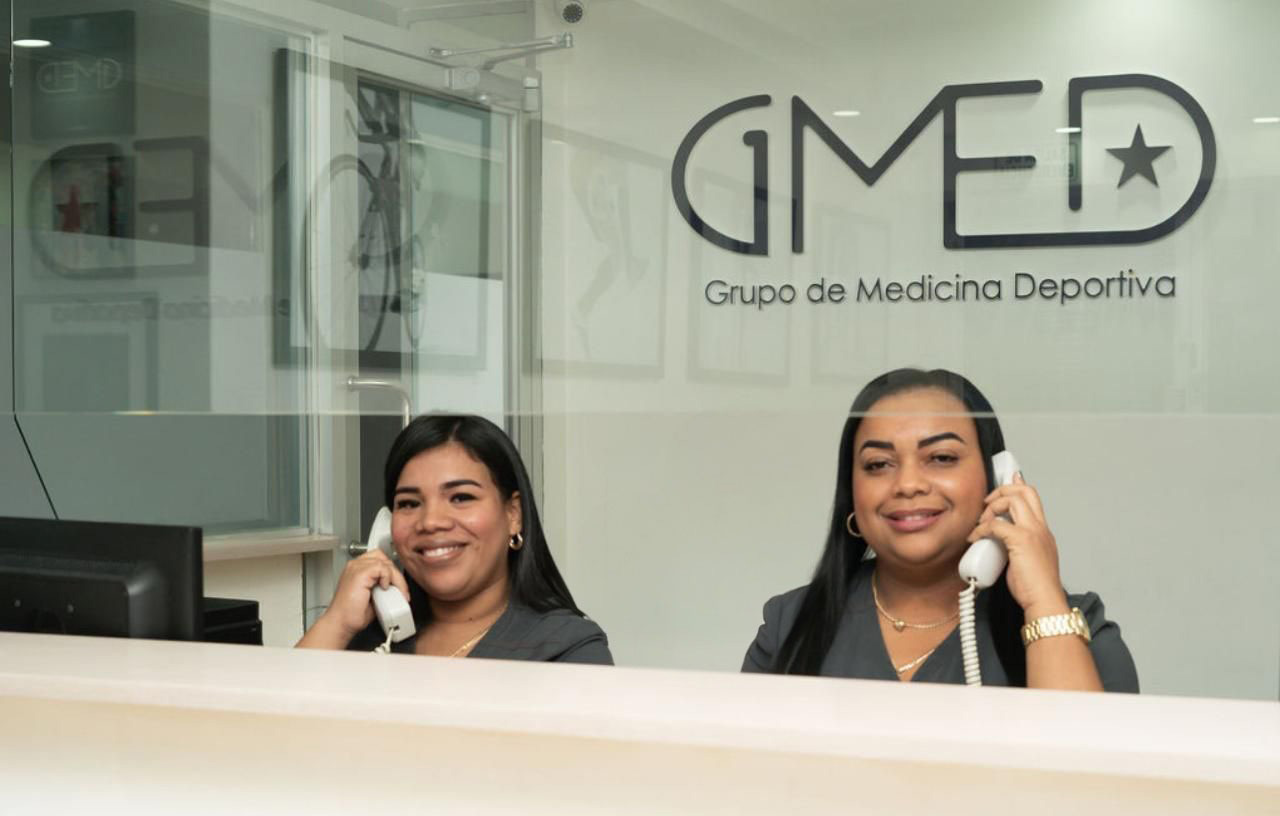 Dos mujeres uniformadas atienden en la recepción de GMEDSport, un centro de medicina deportiva, con sonrisa y atención telefónica. Detrás, un muro de cristal con el logotipo y texto de especialistas en lesiones y ortopedia deportiva.