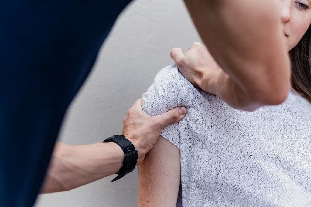 Una persona con camisa blanca recibe fisioterapia en el hombro, mientras otra persona, con reloj negro, le aplica un masaje o análisis, capturando un momento de atención, apoyo y cuidado en un entorno tranquilo.