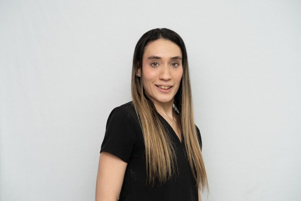 Una persona de pelo castaño, largo y liso, con camisa negra, de pie frente a un fondo liso gris claro, sonríe suavemente a la cámara y encarna su pasión por la nutrición y la vida sana.
